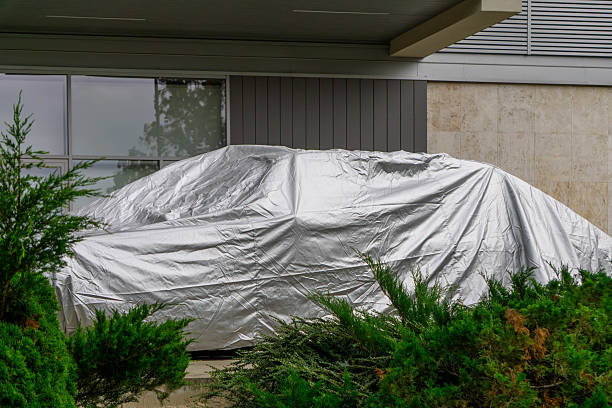 istock car covered with a silver tarp parked under a modern building overhang, surrounded by green shrubs. 2228451549