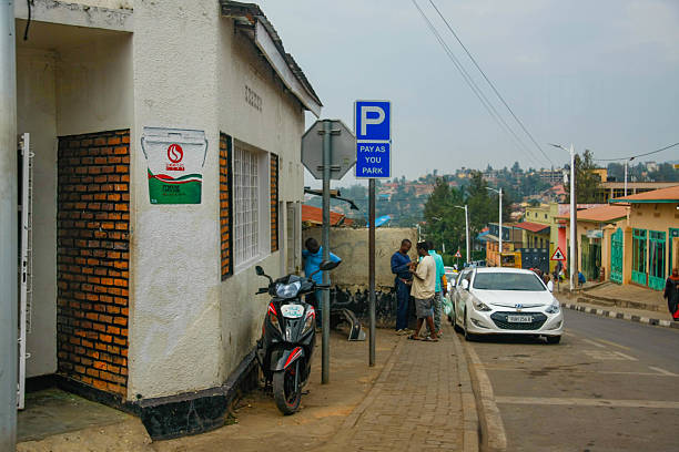 A street corner in the Nyamirambo neighborhood of Kigali, Rwanda, features a prominent blue and white 'P' sign indicating a 'Pay as you Park' zone. A black and red scooter is parked near the sign. Several men are gathered on the sidewalk in front of a light-colored building with a partially exposed brick wall and a sign for 'SIMBA' brand lubricant. A white car is parked along the street, and in the background, colorful buildings and the rolling hills of Kigali are visible under a cloudy sky. This image captures a typical scene of urban life in this vibrant district of Rwanda's capital city.