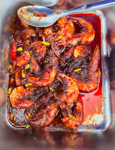 Fresh prawns cooked in a rich, spicy red sambal sauce, garnished with small green bean slices, served in a metal container with a spoon.