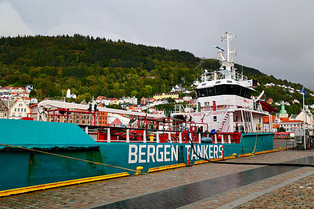 Bergen, Norway - 10 September 2024: A commercial Bergen Tankers vessel docked at the harbor in Bergen, Norway. The ship sits against a picturesque backdrop of colorful hillside homes and dense green forest, characteristic of Western Norway. The waterfront scene highlights the blend of industrial maritime activity with the charming architecture and natural beauty of the region, under a cloudy sky.
