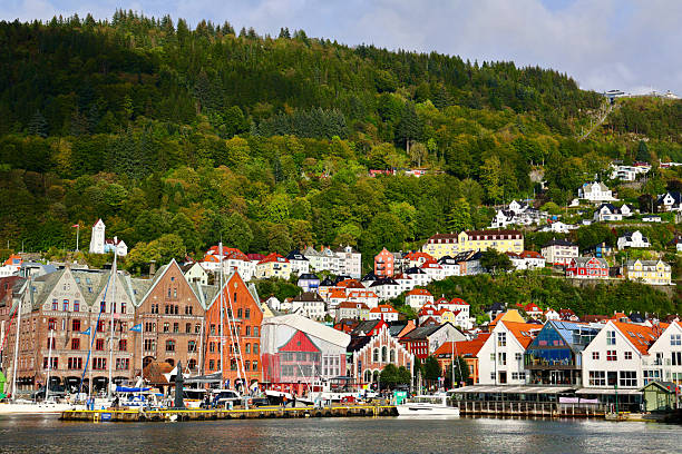 Bergen, Norway - 10 September 2024: A scenic view of Bergen, Norway, showcasing its iconic waterfront with vibrant wooden buildings and a classic ship docked at the harbor. The hillside is covered with charming houses nestled in lush green forest, illustrating the harmony between traditional Norwegian architecture and natural beauty. This image captures the serene coastal atmosphere of one of Scandinavia’s most picturesque cities.