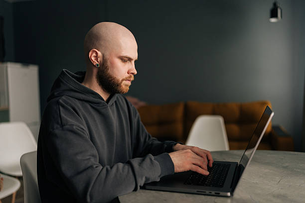 istock Focused bald male programmer with beard and hoodie working typing on laptop computer sitting at table at modern home office, developing software solutions 2228484778