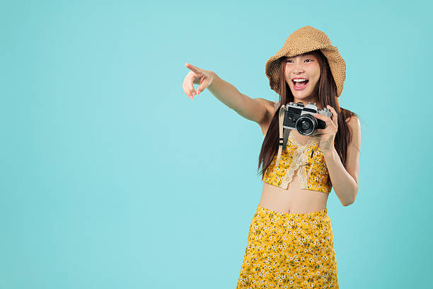 Happy travel tourist asian woman smile vacation, holding camera, a young cheerful tourist points to empty space enjoying her summer holiday in floral outfit, headshots isolated studio blue background