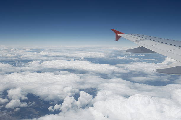 A peaceful in-flight view from a passenger seat showing an aircraft wing soaring above soft clouds under a bright blue sky.