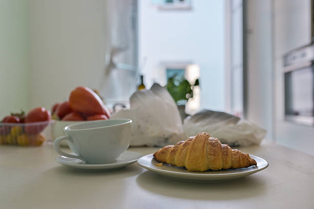 A cozy kitchen scene featuring a croissant on a plate with a coffee cup, fresh produce in the background, illuminated by soft natural morning light from a window.