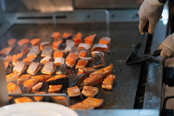 Cubes of salmon sizzling on a flat-top grill, with a gloved chef using a spatula to turn the pieces.