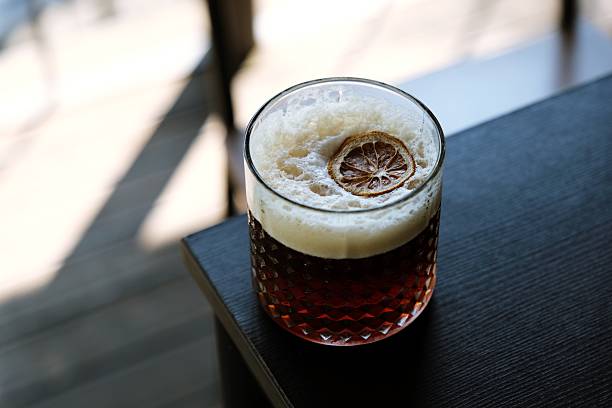 Glass of foamy espresso tonic placed on wooden table with diagonal shadows from sunlight. Warm tones and textured crema visible.