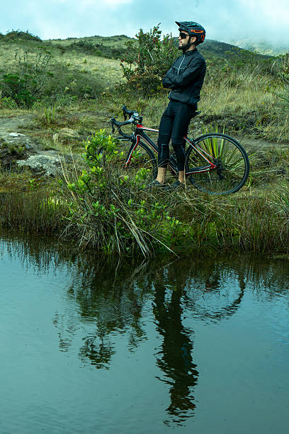 Young cyclist in black windbreaker jacket, protective helmet and glasses, taking a break in mountains and lake after riding a bike