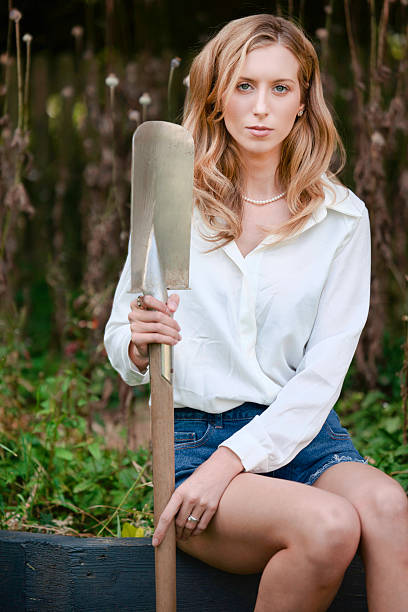 Lady wearing shorts and a blouse and holding a garden spade.