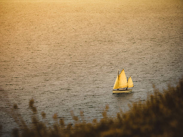 An atmospheric shot of a sailboat on the sea, lit by the warm, golden light of the setting sun in Dorset. The composition focuses on the small boat, which becomes the central point of a vast, tranquil body of water. The depth of field is shallow, with a gently blurred foreground, which enhances the sense of perspective and intimacy of the scene. Captured with an OM-1 Mark II and a 100-400mm telephoto lens. Ideal for themes of travel, peace, dreams, adventure, as well as for marketing materials for the tourism industry and lifestyle publications.