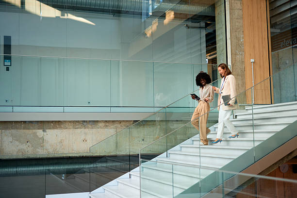 Two smiling businesswomen walking down the stairs of a modern office building, discussing work while holding digital tablets and laptops, embodying collaboration and teamwork