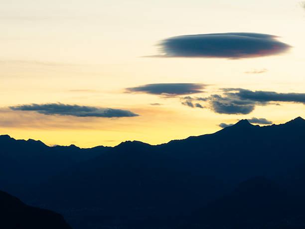 A dramatic mountain skyline is silhouetted against a glowing sunset in the Lombardy Alps of Northern Italy. The jagged peaks form a dark, majestic contrast to the vibrant orange and pink hues of the evening sky, creating a tranquil and timeless alpine landscape. A peaceful end to a day in the high mountains.