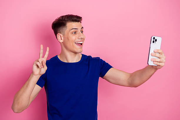 istock Young man in a blue shirt taking a selfie using a smartphone while making a peace sign on a vibrant pink background 2228535951