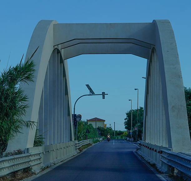 The Mussolini-era concrete arch bridge with its symmetrical pattern and open road is a minimalist perspective under a blue sky, ideal for themes related to history, infrastructure or architecture.