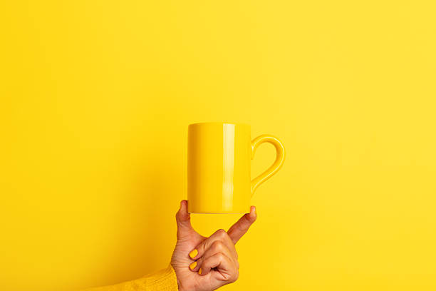 istock Hand in yellow sweater balancing a plain yellow ceramic mug on fingertips against a bright yellow background. 2228538256