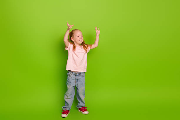 istock Smiling young girl raising her hands with enthusiasm while standing against a bright green background in casual attire 2228538799