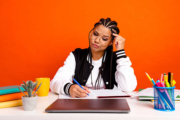 A young female student with braid hairstyle sits at a desk with stationery and laptop, concentrating on her studies