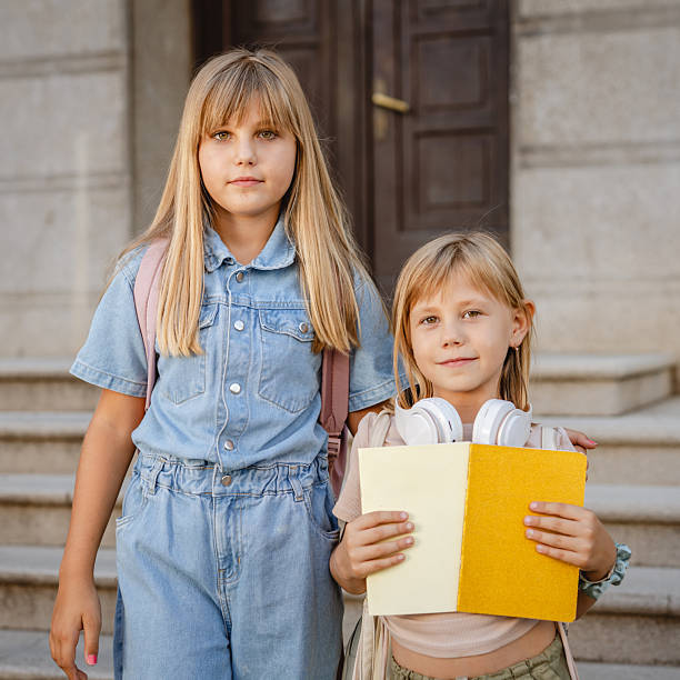 Two young schoolgirls are standing together on the steps of their school, one holding a book and headphones, both wearing backpacks and ready to start their school day