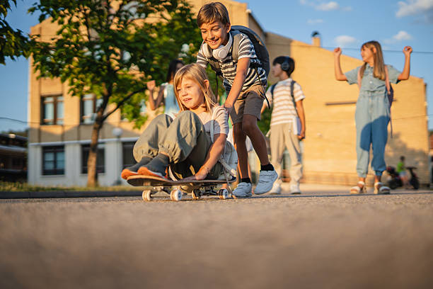Schoolboy pushing his classmate sitting on a skateboard after school, while other students walk and play in the background, enjoying carefree moments of friendship and fun