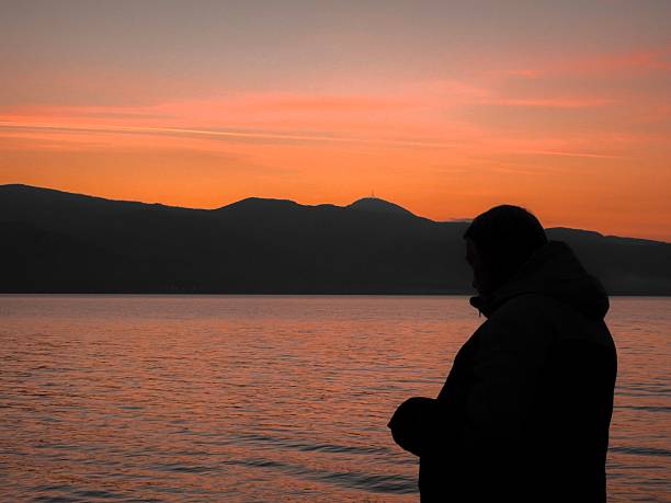 Silhouette of a person gazing at a serene sunset over a calm lake with distant mountains in the background.