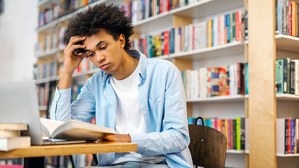 istock Tired black student guy sitting in campus library at table, reading book, preparing to lecture or exam, free space. Knowledge and learning concept 2228553106