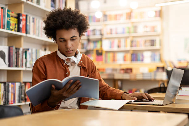istock Concentrated black guy engrossed in study, reading book and using laptop, sitting in university library, preparing for lecture or tests 2228553186
