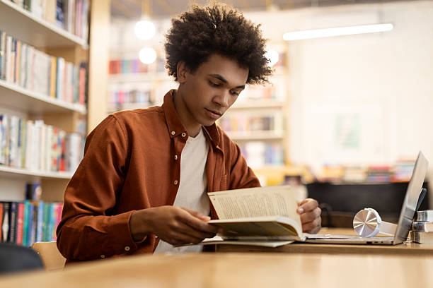 istock Studious African American guy surrounded by books, focuses on educational reading in college library setting, sitting at desk with laptop 2228554163