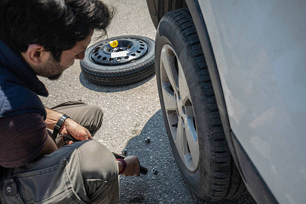 A man is using a lug wrench to remove the wheel bolts on a car while changing a flat tire during a roadside emergency. The spare tire is visible in the background, illustrating an everyday situation involving vehicle maintenance, travel safety, and emergency preparedness. This image is ideal for topics such as car trouble, auto repair, DIY maintenance, and road trip safety