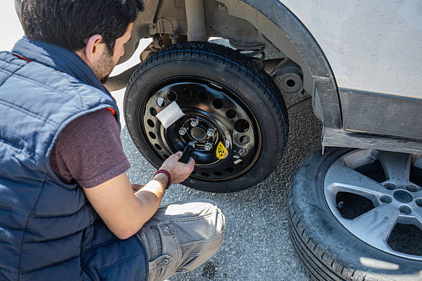 A man wearing a blue vest and casual clothes is seen from behind as he replaces a flat tire with a compact spare wheel on his car, parked on the roadside. Essential tools and a yellow emergency kit are visible in the background, emphasizing roadside emergency, travel safety, and self-reliance. The image highlights do-it-yourself car maintenance, tire changing skills, and handling unexpected breakdowns during a journey