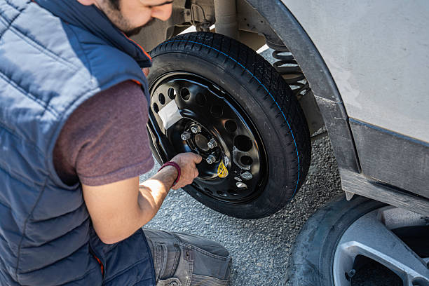 A young man in casual clothing is changing a flat tire with a compact spare wheel on the side of the road after experiencing a car breakdown. He is kneeling and using a lug wrench to secure the spare tire while the damaged tire lies nearby. The image illustrates roadside emergency repair, vehicle maintenance, and travel safety, highlighting self-reliance and practical problem-solving skills during unexpected situations on a journey