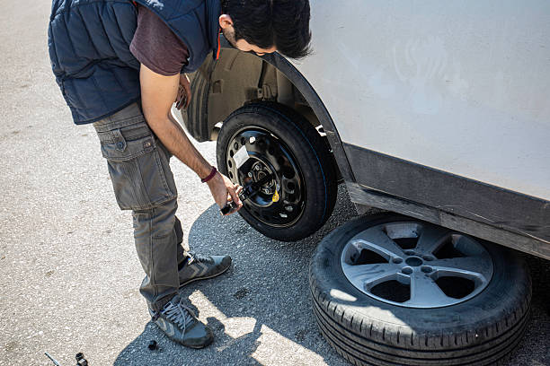 A man in casual clothing is installing a compact spare wheel on his car after a flat tire incident on the roadside. The original flat tire is placed on the ground nearby while he uses a hand tool to securely tighten the bolts. This image highlights car maintenance, roadside emergencies, DIY repair skills, and personal responsibility during travel. Ideal for themes about auto repair, flat tire solutions, travel safety, and emergency preparedness