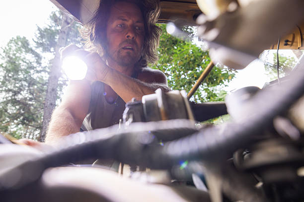 Man inspects a car engine using a flashlight in an outdoor wooded setting. Hands-on automotive repair in natural light, captured from under the hood.