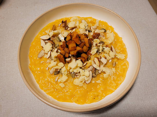 A beautifully plated dish of creamy pumpkin risotto topped with roasted squash cubes, crumbled cheese, sliced almonds, and pumpkin seeds. Served in a ceramic bowl on a neutral tablecloth, this vibrant meal captures the essence of gourmet seasonal cuisine. Ideal for illustrating concepts of autumn food, vegetarian meals, comfort dining, and fine homemade cooking.