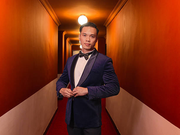 well-groomed Asian man in a dark blue velvet blazer and bow tie stands in a warmly lit hallway, buttoning his jacket with a composed, confident expression. The symmetrical lighting and clean lines create a cinematic mood, blending classic elegance with modern masculinity.