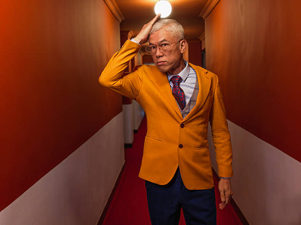 Mature Asian man in a yellow blazer adjusting his hair while walking through a warmly lit hotel corridor. Fashion-forward concept with expressive body language, stylish wardrobe, and a confident attitude.