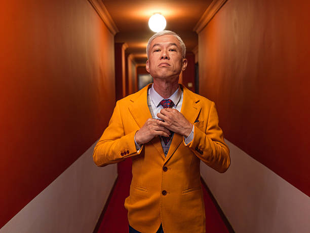 Stylish elderly Asian man in a yellow blazer standing confidently in a modern hotel corridor, adjusting his tie. Shot indoors with dramatic lighting and strong leading lines.