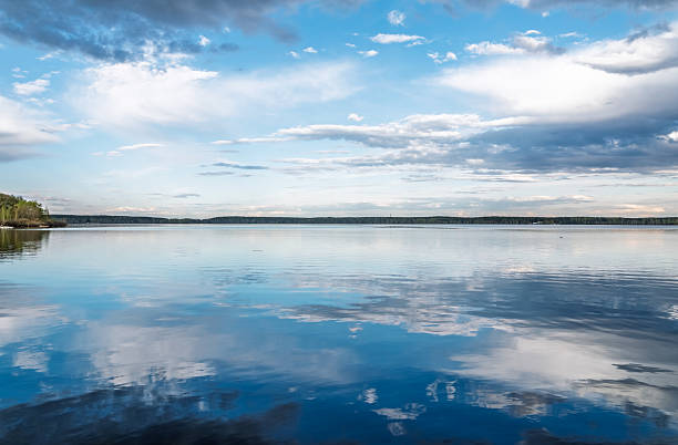 Summers day in Lake Saimaa. Shore showing under tall birch treeon the left side.. Lake surface calm reflecting blue sky with white clouds. Thin section of woods showing in the horizon.