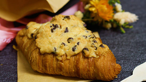 Chocolate chip crookie cookie crowning flaky croissant, resting on parchment with rich dark backdrop showcasing decadent bakery fusion