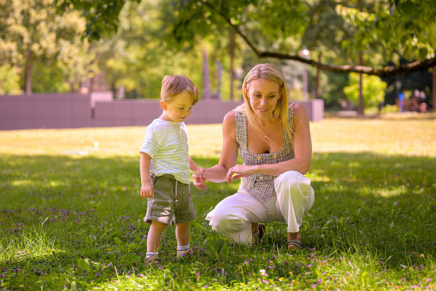 A blonde woman crouches beside a young boy, gently holding his hand as they explore the grassy park together.