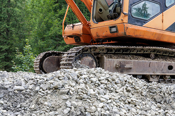 A detailed shot of a large yellow excavator's tracks on a heap of gravel and dirt, with a forest in the background.