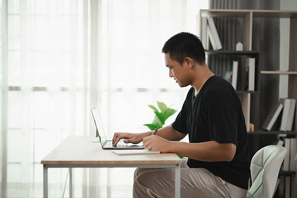 istock Young man working on laptop at home office desk, focused on screen with modern decor and natural light from large windows 2228589758