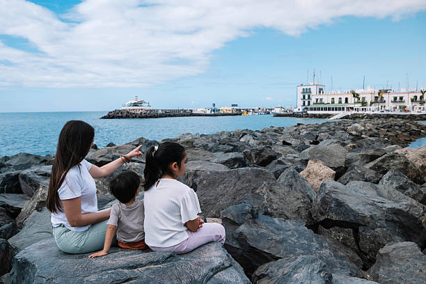 A woman points to a boat in the water. A group of children sit on a rock near the water