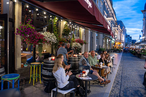 A lively evening atmosphere on a cobblestone street in Vilnius, Lithuania, featuring outdoor cafes and restaurants with patrons enjoying dinner under warm lighting. The historic architecture, vibrant ambiance, and bustling street life capture the charm of Vilnius' city center during twilight.