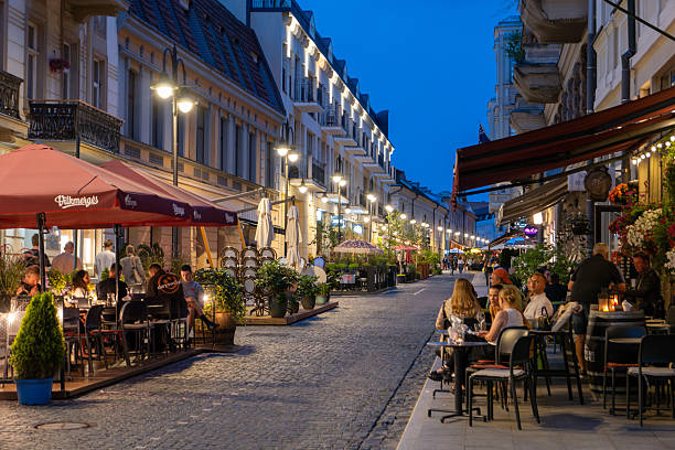 A lively evening atmosphere on a cobblestone street in Vilnius, Lithuania, featuring outdoor cafes and restaurants with patrons enjoying dinner under warm lighting. The historic architecture, vibrant ambiance, and bustling street life capture the charm of Vilnius' city center during twilight.
