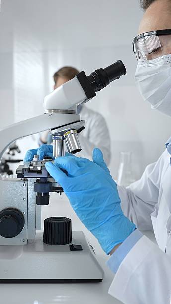 istock Female scientist wearing mask and blue gloves using microscope doing scientific research in modern laboratory with colleague working in background. Medicine, healthcare and science concept 2228595669