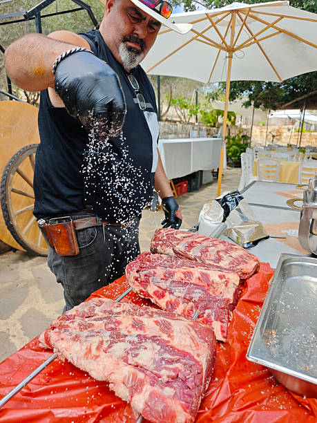 A rugged man in a cowboy hat and black gloves skillfully sprinkles coarse salt over several large slabs of raw ribeye steak skewered and ready for open-fire grilling. The chef exudes confidence and craftsmanship, surrounded by an outdoor rustic setting with umbrellas, wood, and metal elements. This image captures the raw essence of traditional meat preparation, ideal for themes of culinary art, barbecue culture, or outdoor cooking events.