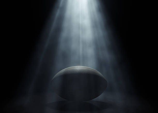 A generic rugby sports ball being lit by a single ethereal light rays on a dark black background - 3D render