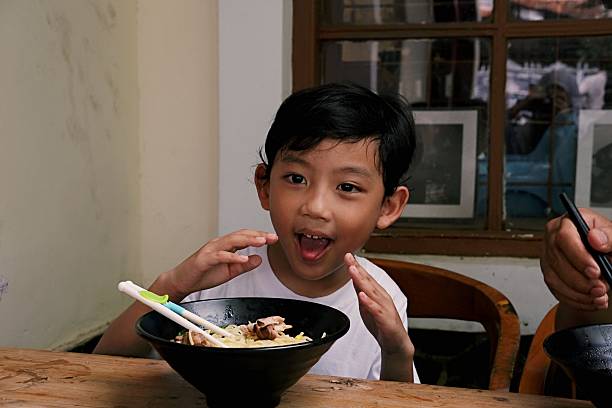 Cheerful young boy using chopsticks while eating ramen noodles at a restaurant table, full of joy.