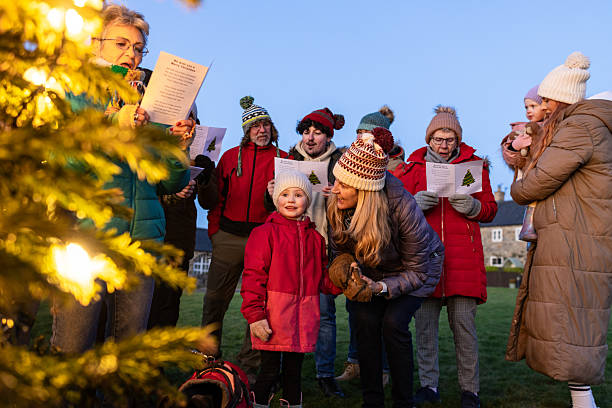 A wide shot of a group of people standing together outside while singing Christmas carols. They are all standing next to a large Christmas tree, which is lit up with warm festive lighting. They are all wearing warm winter clothing with woolly hats. They are holding a booklet with Christmas carol lyrics. Everyone is smiling as they have come together as a community to celebrate Christmas.Videos are available similar to this scenario.