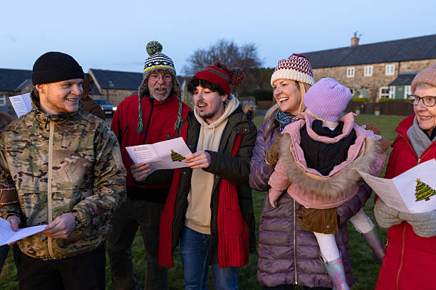 A wide shot of a group of people standing together outside while singing Christmas carols. They are all wearing warm winter clothing with woolly hats. They are holding a booklet with Christmas carol lyrics. Everyone is smiling as they have come together as a community to celebrate Christmas.Videos are available similar to this scenario.
